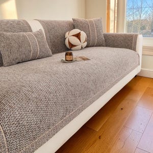 Könnte beinhalten: Ein graues Stoffsofa mit weißen Zierleisten, mit passenden Kissen mit hellrosa Besatz. Ein dekorativer Ball und eine Kerze liegen auf dem Sofa. Das Sofa steht in einem hellen Raum mit Parkettboden.