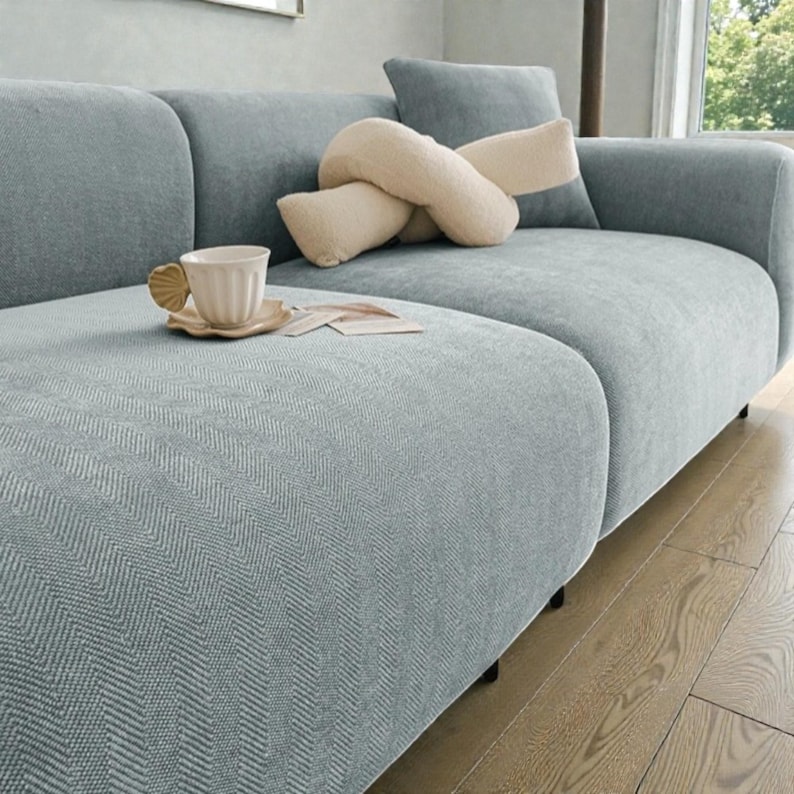 May include: A light blue sofa with a textured herringbone pattern, featuring a cream-colored knot pillow and a fluted ceramic cup with a shell-shaped handle resting on a matching saucer, placed on the seat cushion of the upholstered furniture.