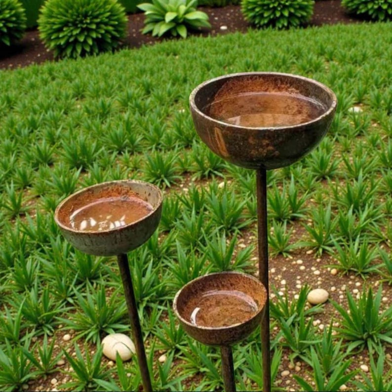 May include: Three rustic metal bird baths of varying heights are set in a lush green garden. The bowls are filled with water, reflecting the sky. The bird baths have a weathered, aged appearance, adding a touch of vintage charm to the outdoor space.