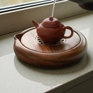 May include: A small, reddish-brown ceramic teapot with a matching lid is placed on a round, wooden tea tray. The tray has a raised edge and a metal grate in the center. Water is being poured into the teapot. The tea set is on a light-colored surface.
