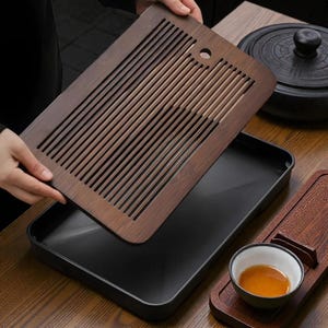Könnte beinhalten: Ein dunkles Holz-Teetablett mit einem Lattenrost wird über einem schwarzen Tablett gehalten. Das Teetablett hat eine dunkelbraune Oberfläche und ein kleines Loch in der Ecke. Eine kleine Teetasse mit Tee steht auf einem Holzuntersetzer.