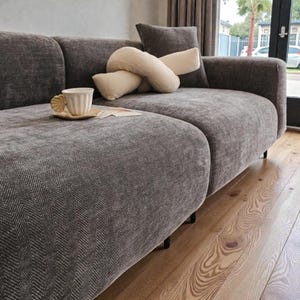 May include: A dark gray sofa with a textured herringbone pattern, featuring a cream-colored knotted throw pillow and a small, light-colored ceramic mug with a shell-shaped handle resting on a matching saucer placed on the seat cushion.