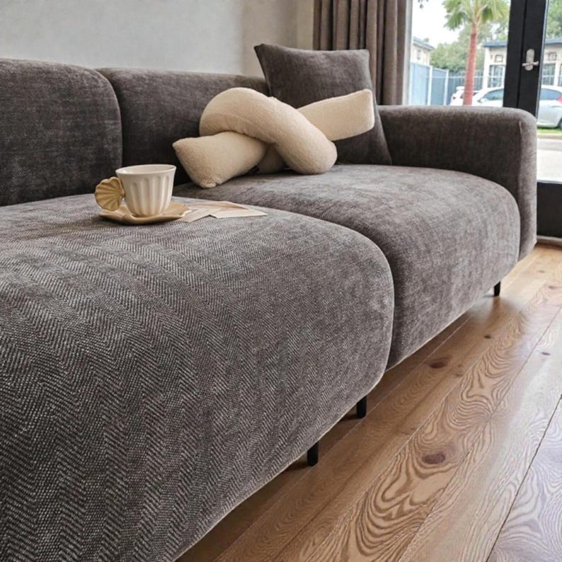 May include: A dark gray sofa with a textured herringbone pattern, featuring a cream-colored knotted throw pillow and a small, light-colored ceramic mug with a shell-shaped handle resting on a matching saucer placed on the seat cushion.
