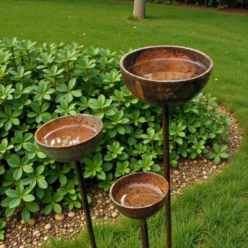 May include: Three rustic, metal bird baths of varying sizes stand in a garden. Each bowl-shaped bath is filled with water and mounted on a slender, dark brown pole. The bird baths are surrounded by green foliage and grass.