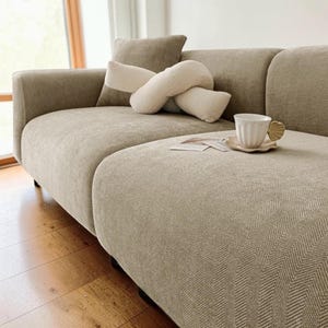 May include: A beige sectional sofa with a patterned cushion and a matching ottoman. Decorative pillows in beige and cream tones rest on the sofa. A white teacup and saucer sit on the ottoman.