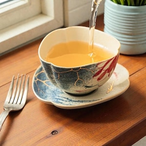 May include: A floral-decorated teacup and saucer set with a blue and white design. The teacup is filled with tea, and a fork rests on the wooden surface. The teacup has a unique shape and is placed on a matching saucer.