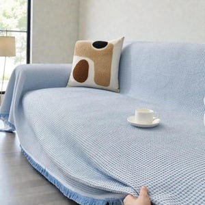 Puede incluir: Una funda de sofá azul claro con un patrón de tejido de gofre y ribete de flecos. Un cojín decorativo con formas abstractas en marrón, negro y beige descansa sobre el sofá. Una taza y un platillo blancos están sobre la funda.