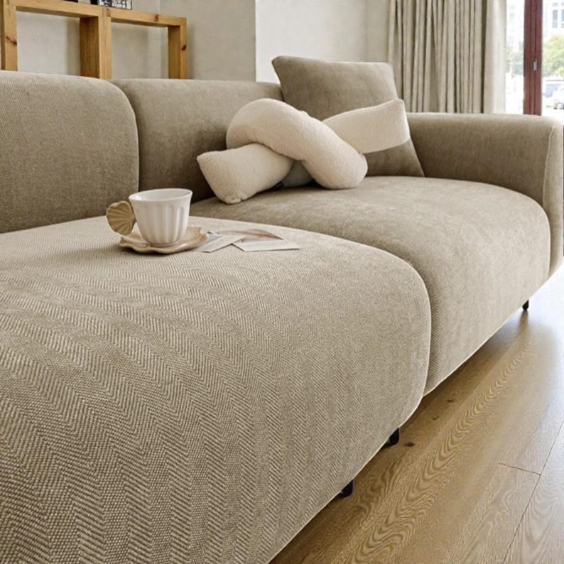 May include: A beige sofa with a subtle herringbone pattern, featuring a white ceramic mug with a shell-shaped handle on a matching saucer, and a cream-colored knotted throw pillow resting on the seat cushion of the upholstered furniture.