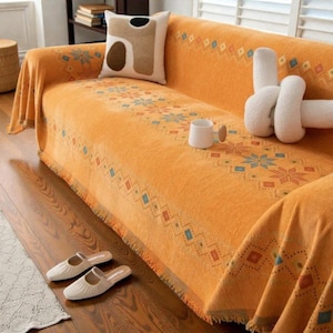 May include: An orange sofa cover with a geometric pattern and fringing. The cover is draped over a sofa, with a decorative cushion and a white mug on top. A pair of cream-coloured slippers are on the wooden floor.