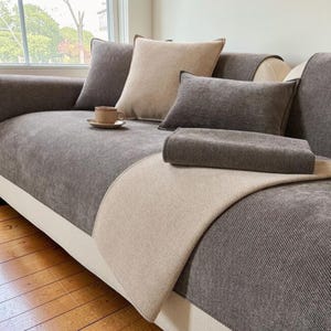 Könnte beinhalten: Ein graues und beiges Sofa mit mehreren Kissen und einer gefalteten Decke. Das Sofa hat eine weiße Basis und Holzböden. Eine Tasse und Untertasse stehen auf dem Sofa. Die Kissen sind in Grau- und Beigetönen gehalten.
