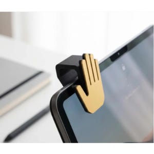 May include: A black and gold hand-shaped tablet camera cover. The cover is attached to the top edge of a tablet, with the gold hand design facing forward. A notebook and stylus are in the background.