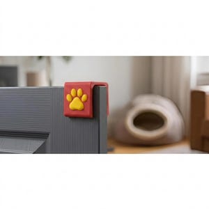 May include: A red door stop with a yellow paw print design. The door stop is attached to a gray door. A pet bed is in the background.