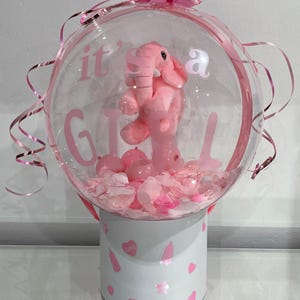 May include: A clear bubble balloon with pink ribbons and a pink bow. Inside, a pink elephant soft toy is surrounded by pink confetti and small balloons. The text "it's a GIRL" is printed on the balloon. The balloon sits on a white box decorated with pink hearts and baby bottle designs.