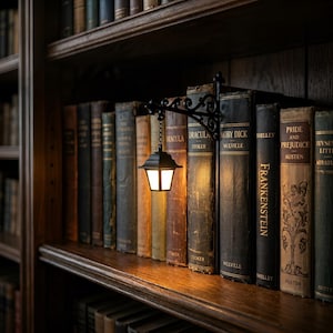May include: A black metal wall-mounted lantern light fixture with an ornate scrollwork bracket, suspended by a small chain between vintage hardcover books on a wooden shelf. The lantern emits a warm, soft glow, illuminating the spines of the books.