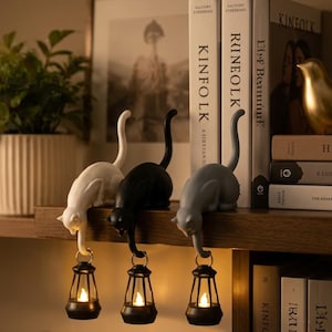 May include: Three cat figurines in white, black, and gray, each perched on a shelf edge. Each cat holds a small black lantern with a glowing light suspended from its tail. The cats are positioned in a row, with their bodies curved downward.