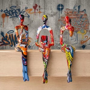 May include: Three abstract human-shaped sculptures sitting in a row. Each figure features a vibrant, multi-colored graffiti-style pattern with bold splashes of red, blue, yellow, and black. The figures have elongated limbs and smooth, glossy surfaces.