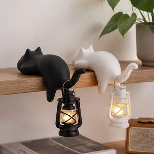 May include: Two cat-shaped figurines, one black and one white, are positioned on a shelf. Each cat has a curved tail that holds a small lantern. The black cat holds a black lantern, and the white cat holds a white lantern with a glowing light.