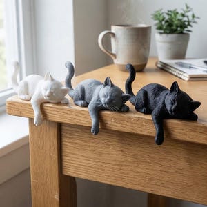 May include: Three small cat figurines in white, grey, and black are positioned along the edge of a wooden surface. Each cat is depicted in a sleeping pose with one front paw hanging over the edge, featuring detailed fur textures and upright tails.