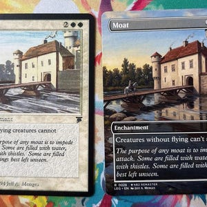 Puede incluir: Dos cartas "Moat" de Magic: The Gathering. Las cartas presentan una ilustración en acuarela de un castillo con un puente sobre un foso. El texto describe el efecto del encantamiento en criaturas no voladoras.