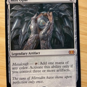 Pode incluir: Uma carta colecionável Mox Opal com uma ilustração metálica de mãos segurando um orbe brilhante. O texto da carta diz Legendary Artifact, Metalcraft e inclui o texto de ambientação The suns of Mirrodin have shone upon perfection only once.