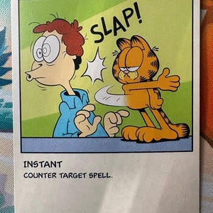 Pode incluir: Uma carta colecionável retangular intitulada COUNTERSPELL com uma ilustração de um gato laranja dando um tapa em um homem de camisa azul. O texto SLAP! aparece acima do gato. A carta inclui o texto INSTANT e COUNTER TARGET SPELL.