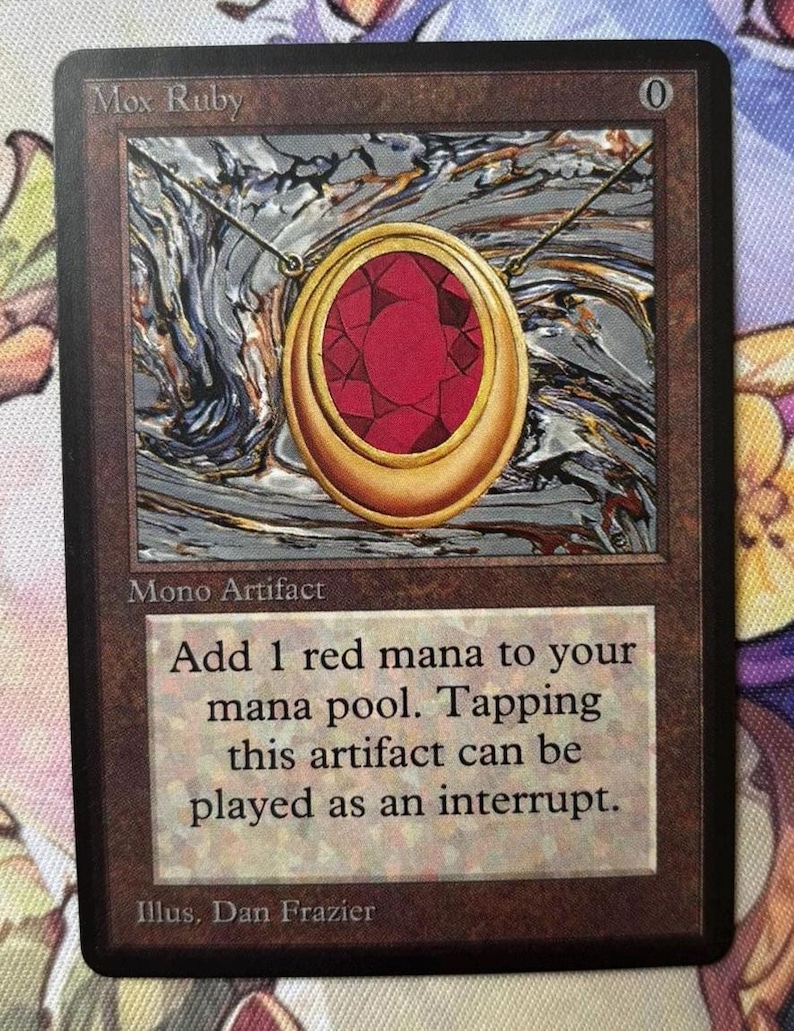 May include: A trading card depicting a gold pendant with a large red gemstone, titled "Mox Ruby." The card includes text: "Add 1 red mana to your mana pool. Tapping this artifact can be played as an interrupt."