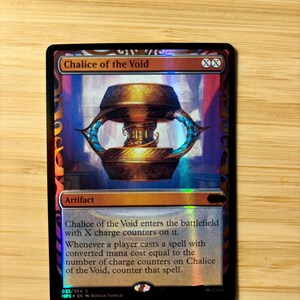 Puede incluir: Una carta coleccionable holográfica de Magic: The Gathering titulada Chalice of the Void. La carta presenta una ilustración de un cáliz de oro metálico, detalles en naranja y azul, y texto negro que detalla la habilidad del artefacto.