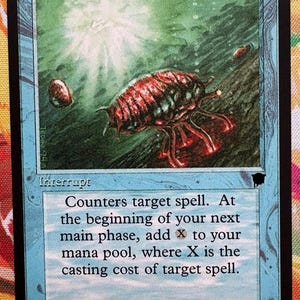 Peut inclure: Une carte à collectionner Mana Drain représentant une créature rouge segmentée semblable à un insecte sous l'eau. La carte a une bordure bleu clair, un encadré noir et le texte : Counters target spell. At the beginning of your next main phase.
