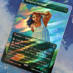 Può includere: Una carta collezionabile olografica di Magic: The Gathering intitolata Delighted Halfling. La carta presenta l'illustrazione di un mezzuomo sorridente con capelli scuri e un vestito chiaro, con testo che descrive le sue abilità come creatura.