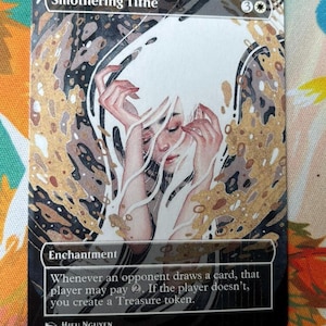 Può includere: Una carta collezionabile Smothering Tithe con l'illustrazione di una figura dai lunghi capelli bianchi e le mani vicino al viso. Il testo recita Enchantment, Whenever an opponent draws a card, that player may pay 2. If the player doesn't, you create a Treasure token. È presente il nome dell'artista Hieu Nguyen.