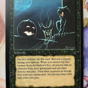 Puede incluir: Una carta de Magic: The Gathering titulada "All Hallow's Eve". La carta presenta una ilustración espeluznante con una calabaza, una figura demoníaca y una criatura felina bajo la luna llena. La carta es de 1994.