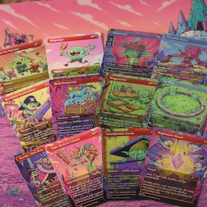 May include: A collection of ten colourful trading cards featuring goblin illustrations and game text, arranged in a fan shape. The cards display various fantasy characters and scenes, resting on a playmat with a pink and purple landscape design.