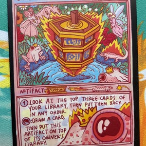 May include: A rectangular trading card titled Sensei's Divining Top featuring a gold spinning top surrounded by flames, a frog, a fairy, and a lizard. The bottom section contains text describing game rules, a large red eye, and a crown. The card is black-bordered.