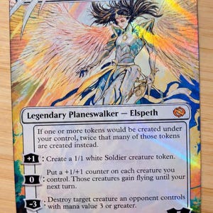 May include: A holographic Magic: The Gathering trading card titled Elspeth, Storm Slayer. The card features a winged figure in armor with flowing hair, surrounded by iridescent colors. It includes game text, ability icons, and the artist name Molly Mendoza.