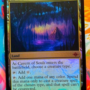 May include: A holographic trading card titled "Cavern of Souls." The card features a mystical cave scene with a pyramid structure. The text includes instructions for gameplay, with the card's type listed as "Land."