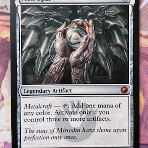 Può includere: Una carta di Magic: The Gathering intitolata "Mox Opal". La carta presenta un'illustrazione dettagliata di mani metalliche che tengono un globo luminoso. Il testo include "Artefatto Leggendario" e le istruzioni del gioco. La firma dell'artista è in basso.