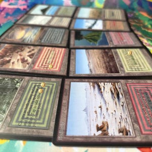 Può includere: Una collezione di carte collezionabili rettangolari disposte in file, ognuna con un'illustrazione paesaggistica nella parte superiore e una casella di testo sotto. Le carte mostrano varie immagini di terreno, montagne e pianure.