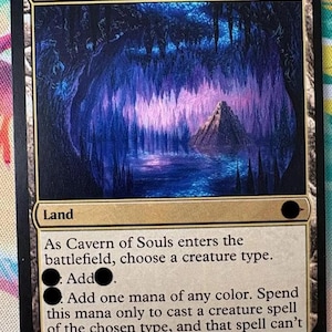 Puede incluir: Una carta coleccionable rectangular titulada Cavern of Souls que presenta una ilustración de una caverna en azul y morado con una estructura piramidal central. El texto incluye reglas de juego, tipo tierra y el nombre Alayna Danner.
