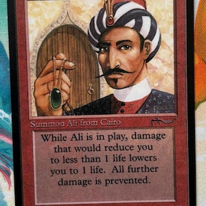 May include: A red Magic: The Gathering trading card titled Ali from Cairo featuring an illustration of a person wearing a black and white striped turban and holding a green gemstone pendant. The card text describes the ability to prevent damage.