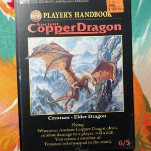 May include: A rectangular trading card with a black border featuring the text Player's Handbook and Ancient Copper Dragon. The central illustration depicts a copper-colored dragon flying over snowy mountains. The card includes game text and a 6/5 rating.