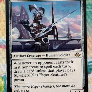 May include: A Magic: The Gathering card titled "Esper Sentinel." The card features a futuristic warrior holding a sword, set against a backdrop of a futuristic city. The card text describes the creature's abilities.