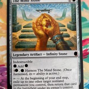 May include: A rectangular trading card titled The Mind Stone, featuring an illustration of golden hands holding a glowing yellow orb. The card text includes Legendary Artifact - Infinity Stone, Indestructible, and specific game abilities. The border is black.