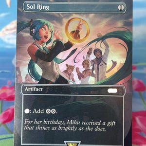 May include: A rectangular trading card titled Sol Ring featuring an illustration of a teal-haired character holding a golden ring. The card includes the text Artifact, Add two colorless mana, and a flavor text about Miku's birthday.