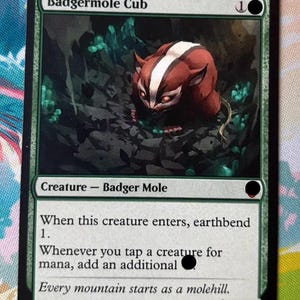 Puede incluir: Una carta coleccionable rectangular titulada Badgermole Cub que muestra una criatura marrón y blanca en un entorno rocoso oscuro con cristales verdes. El texto incluye el tipo de criatura Badger Mole, reglas de juego y el texto de ambientación Every mountain starts as a molehill.