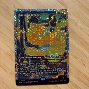 May include: A holographic Magic: The Gathering trading card titled Enduring Tenacity. The card features a yellow, glowing snake-like creature illustration, black text boxes, and a 4/3 power and toughness rating, all set against a shimmering, reflective foil background.
