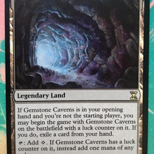 以下が含まれることがあります： Gemstone Cavernsというタイトルの長方形のMagic: The Gatheringトレーディングカード。カードには暗く輝く洞窟の内部のイラストが描かれています。テキストには、特定のゲームルールとマナ能力を持つ伝説の土地としてカードが説明されています。