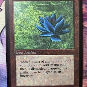 Peut inclure: Carte de jeu avec une fleur de lotus bleue sur fond vert. La carte est intitulée "Black Lotus" et est un Mono Artifact. Le texte décrit sa fonction dans le jeu. Illustration de Christopher Rush.