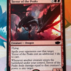 Peut inclure: Une carte à collectionner Terror of the Peaks représentant un dragon noir aux accents rouges sur un fond sombre et tourbillonnant. Le texte de la carte indique le nom, le coût, le type de créature, la capacité de vol et les règles de jeu, 5/4.