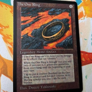 Puede incluir: Una carta coleccionable de Magic: The Gathering titulada The One Ring, que muestra un anillo dorado sobre roca volcánica oscura y lava. El texto de la carta describe sus habilidades de artefacto legendario y el nombre del ilustrador.