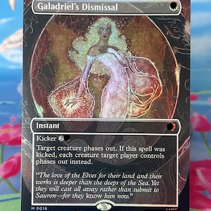 Puede incluir: Una carta coleccionable de Magic: The Gathering titulada Galadriel's Dismissal, con la ilustración de una figura de largo cabello rubio y vestido blanco. El texto de la carta incluye las reglas del hechizo instantáneo y una cita.